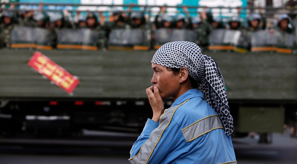 Mulher uigure na beira da estrada enquanto polícia paramilitar chinesa passa dentro de caminhão em rua de Urumqi, na região de Xinjiang, na China