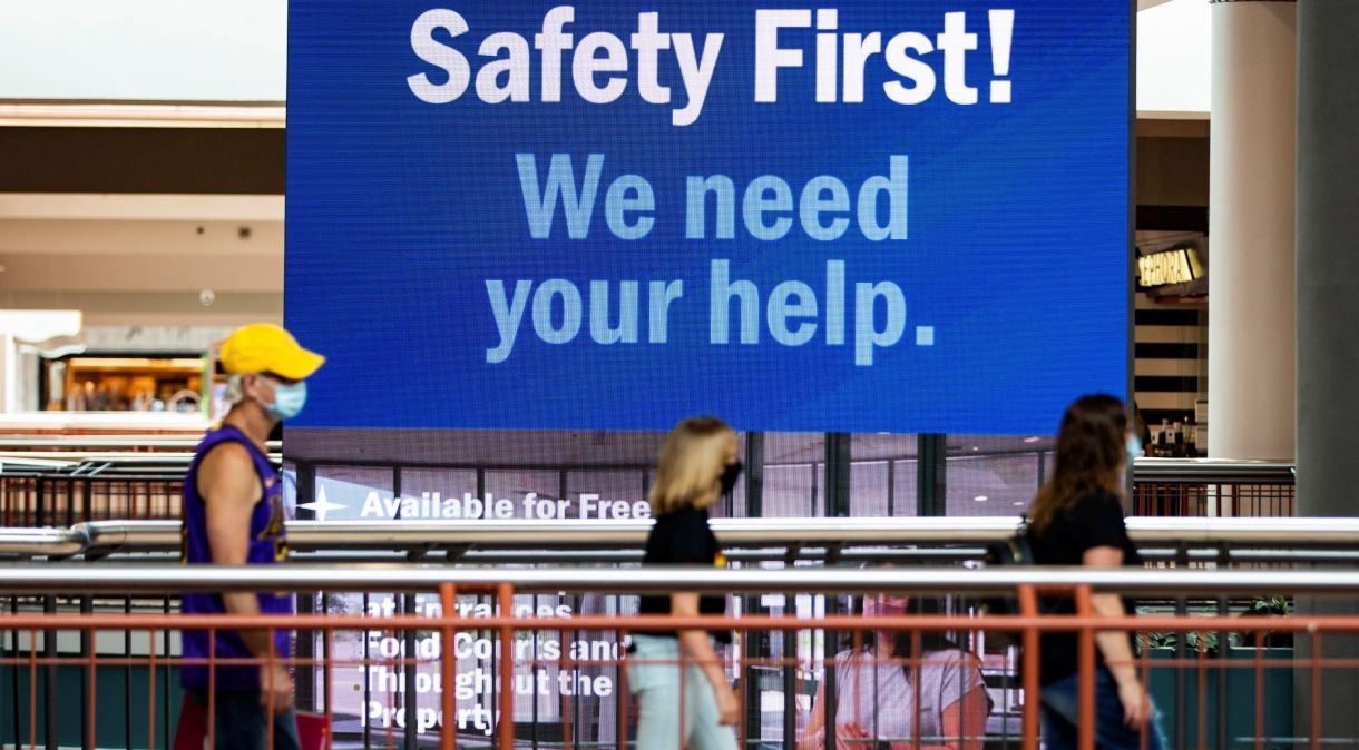 Pessoas caminham de máscara em shopping no estado de Nova York; atrás, a placa diz "Segurança em primeiro lugar! Precisamos de sua ajuda"