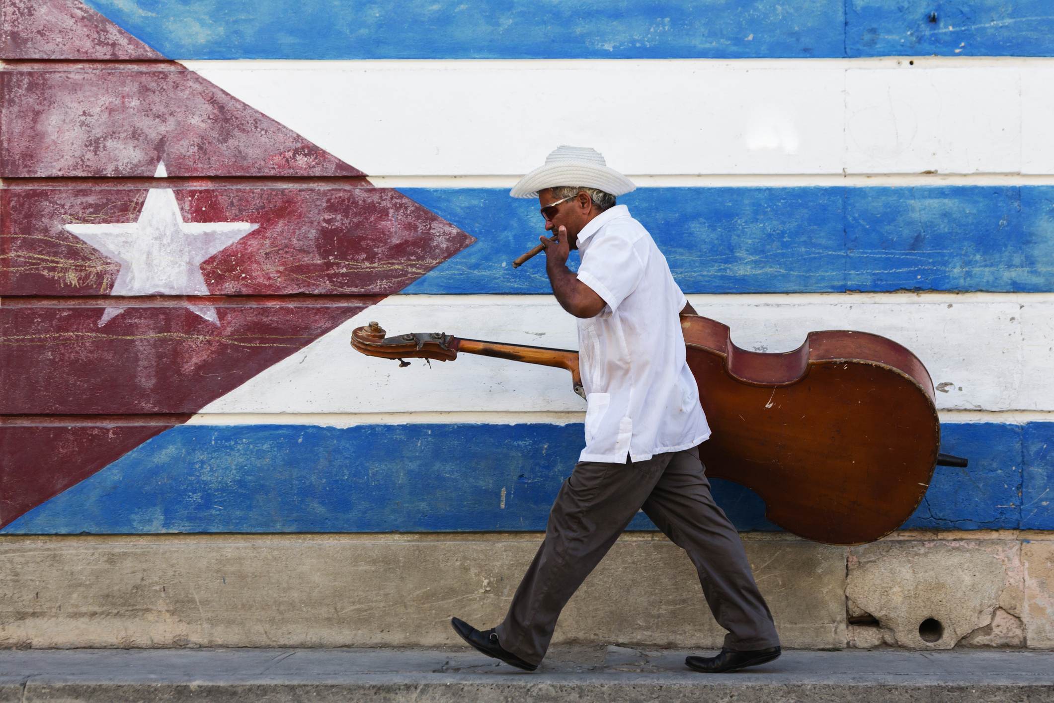 Músico cubano caminha com seu instrumento musical diante de um mural com a bande