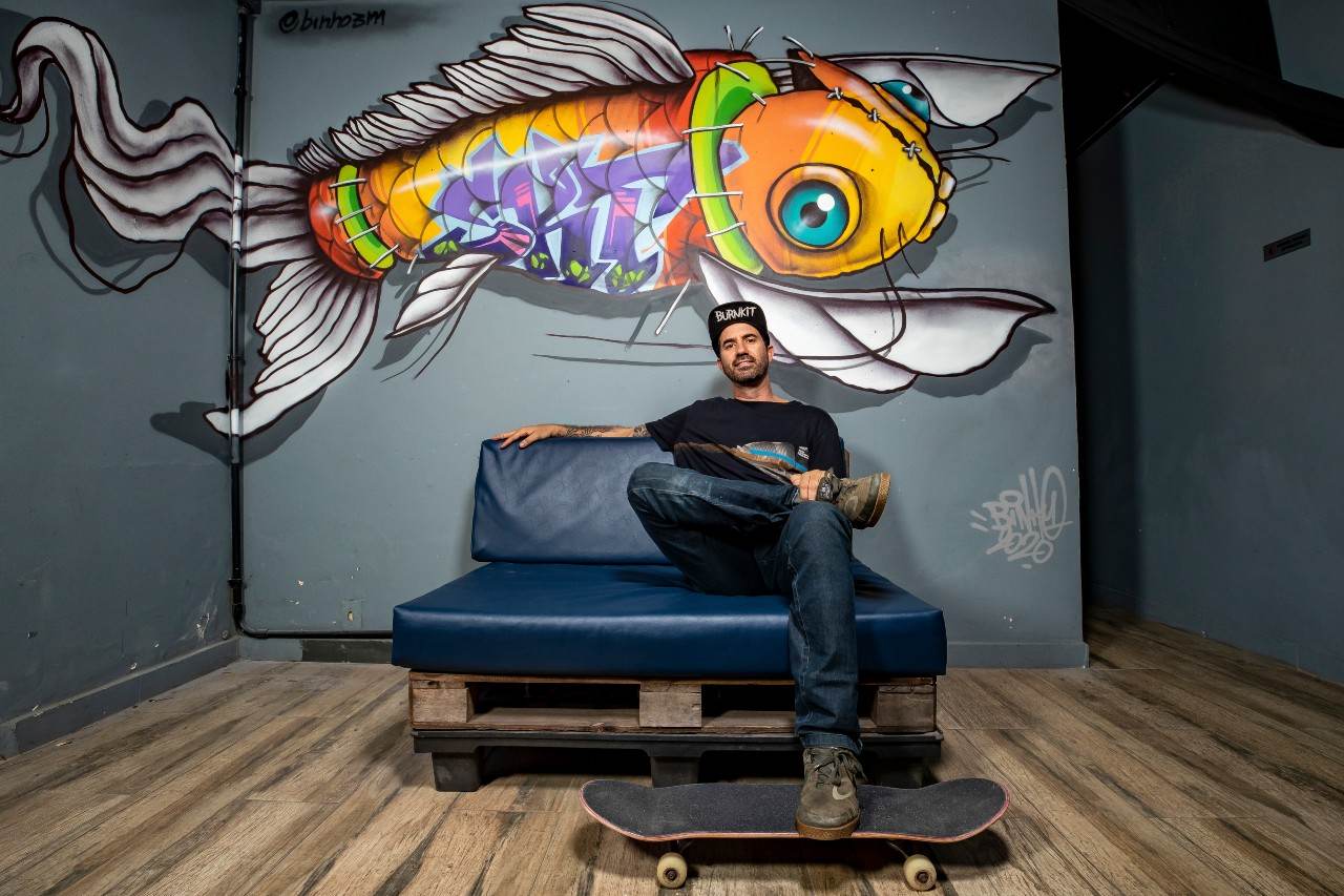 Bob Burnquist sentado em um sofá em frente a um grafite com o skate no pé