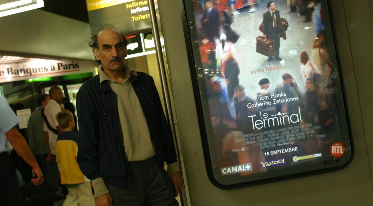 Mehran Karimi Nasseri ao lado de um cartaz do filme de Steven Spielberg "O Terminal", que foi vagamente baseado em sua vida encalhado no Aeroporto Charles de Gaulle