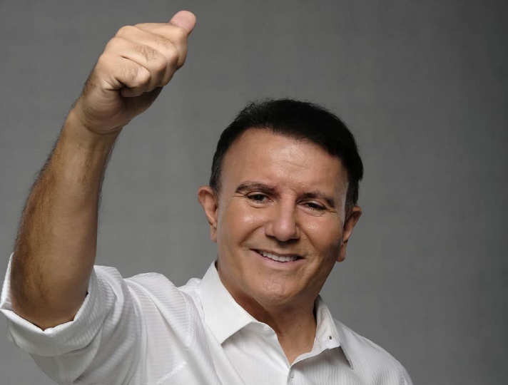 Foto mostra Eduardo Siqueira Campos com um braço para cima. Ele veste uma camisa branca e sorri. Atrás, há um fundo cinza. 