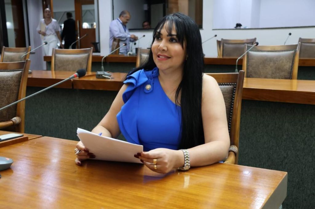 Foto mostra Janad Valcari sentada em uma mesa na Assembleia Legislativa de Tocantins. Ela veste uma roupa azul, tem cabelo preto e usa maquiagem.