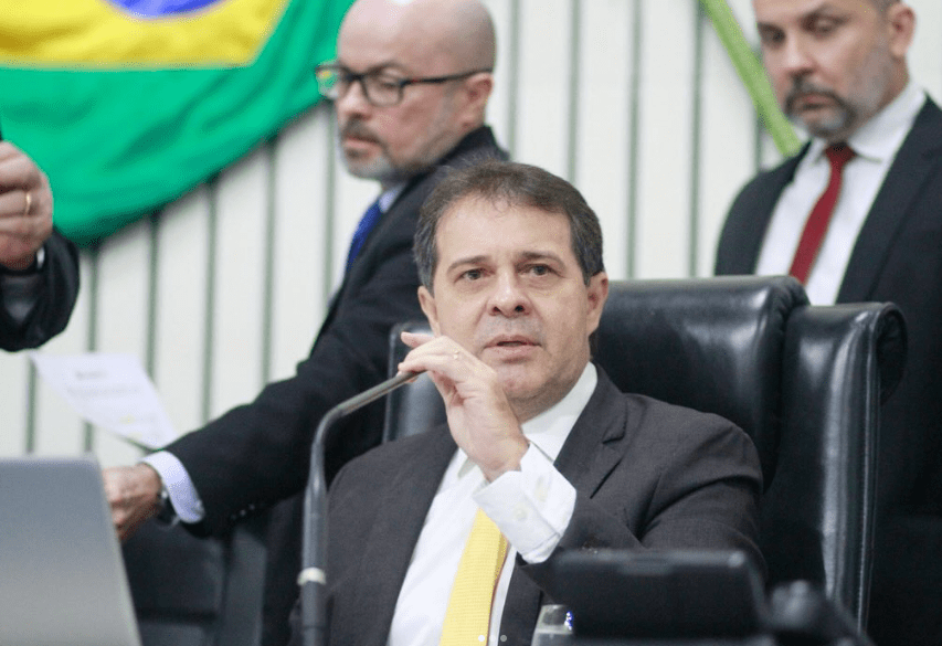 Na foto, Evandro Leitão aparece segurando um microfone. Ele veste um terno preto, gravata amarela e camisa branca. 