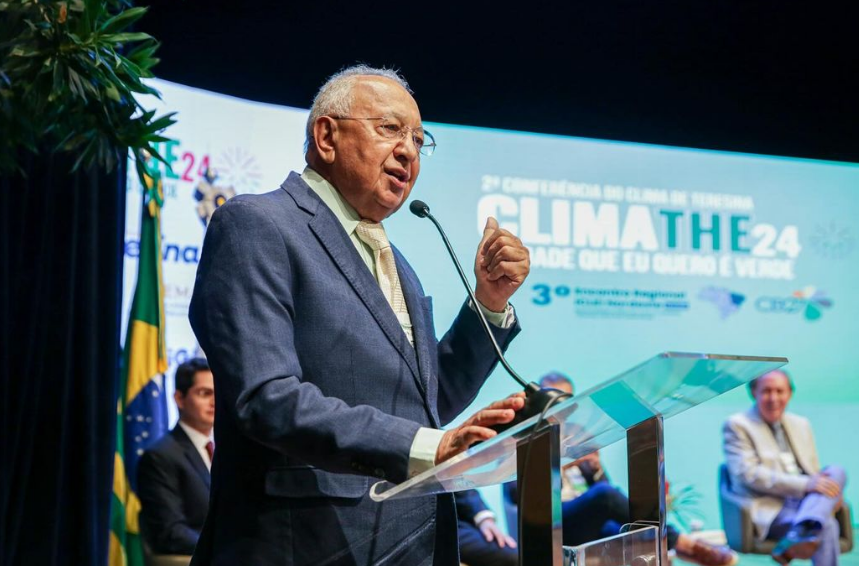 Foto mostra Dr. Pessoa em um evento. Ele tem cabelo grisalho, usa óculos, terno azul escuro, camisa verde e gravata amarela