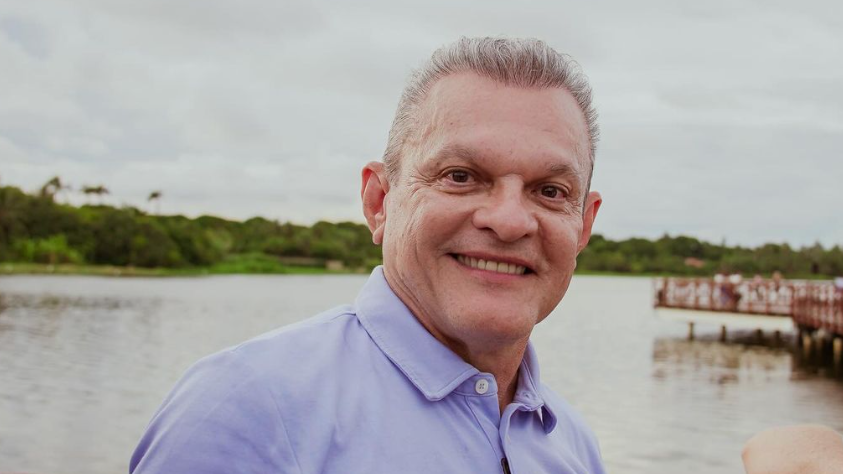 José Sarto aparece com uma camisa lilás e sorrindo. Atrás, há a um rio.