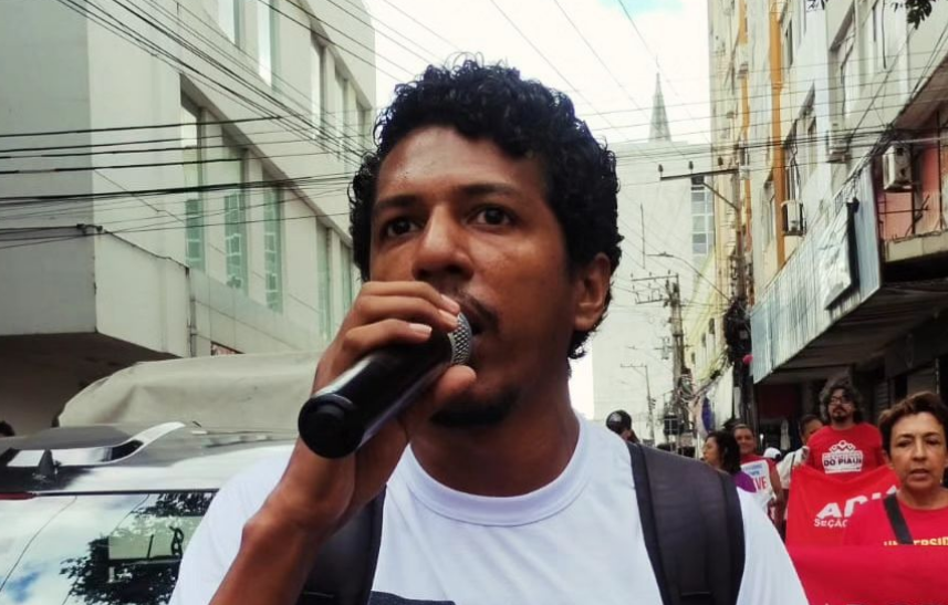 Foto mostra Santiago Belizário (Unidade Popular) com um microfone na rua. Ele é negro, tem bigode e cabelo pretos. Usa uma blusa branca estampada com uma ilustração preta. 