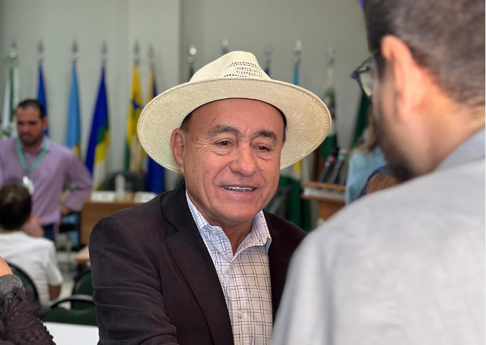 Tião Bocalom veste terno preto, camisa quadriculada e chapéu de palha. Ele é idoso e sorri. 